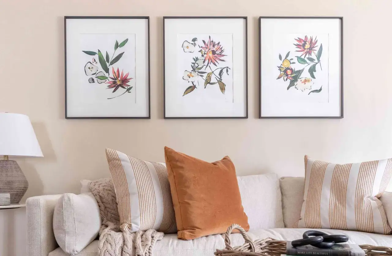 Disposition 3 cadres avec des aquarelles de fleurs et de feuilles vertes, posés au-dessus d'un canapé confortable avec des coussins texturés.
