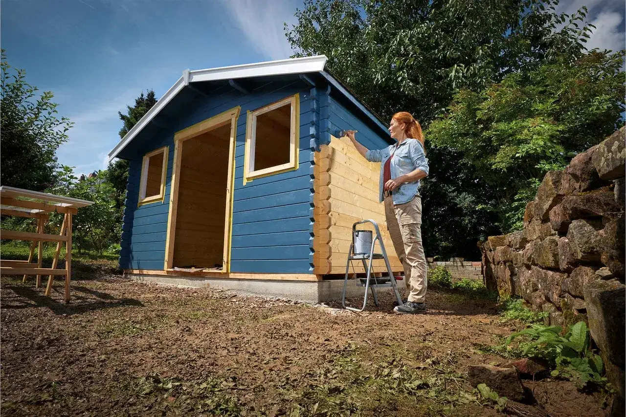 Frau streicht ihr Gartenhaus blau an. Ein Teil ist bereits gestrichen, der andere Teil ist noch naturbelassen.