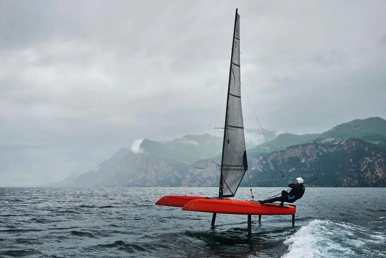 Un voilier orange futuriste fend les vagues, ses foils hors de l'eau. Le marin, penché, semble naviguer vers de nouveaux horizons, porté par les **sails of change**.
