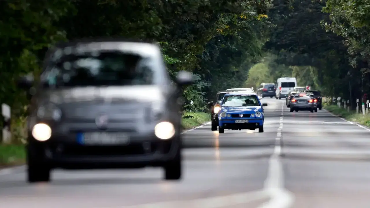 Autos auf einer Stra&szlig;e, die von B&auml;umen ges&auml;umt ist. Ein graues Auto ist im Vordergrund unscharf, ein blaues Auto f&auml;hrt auf der Gegenfahrbahn. Der halber tacho ist erreicht.