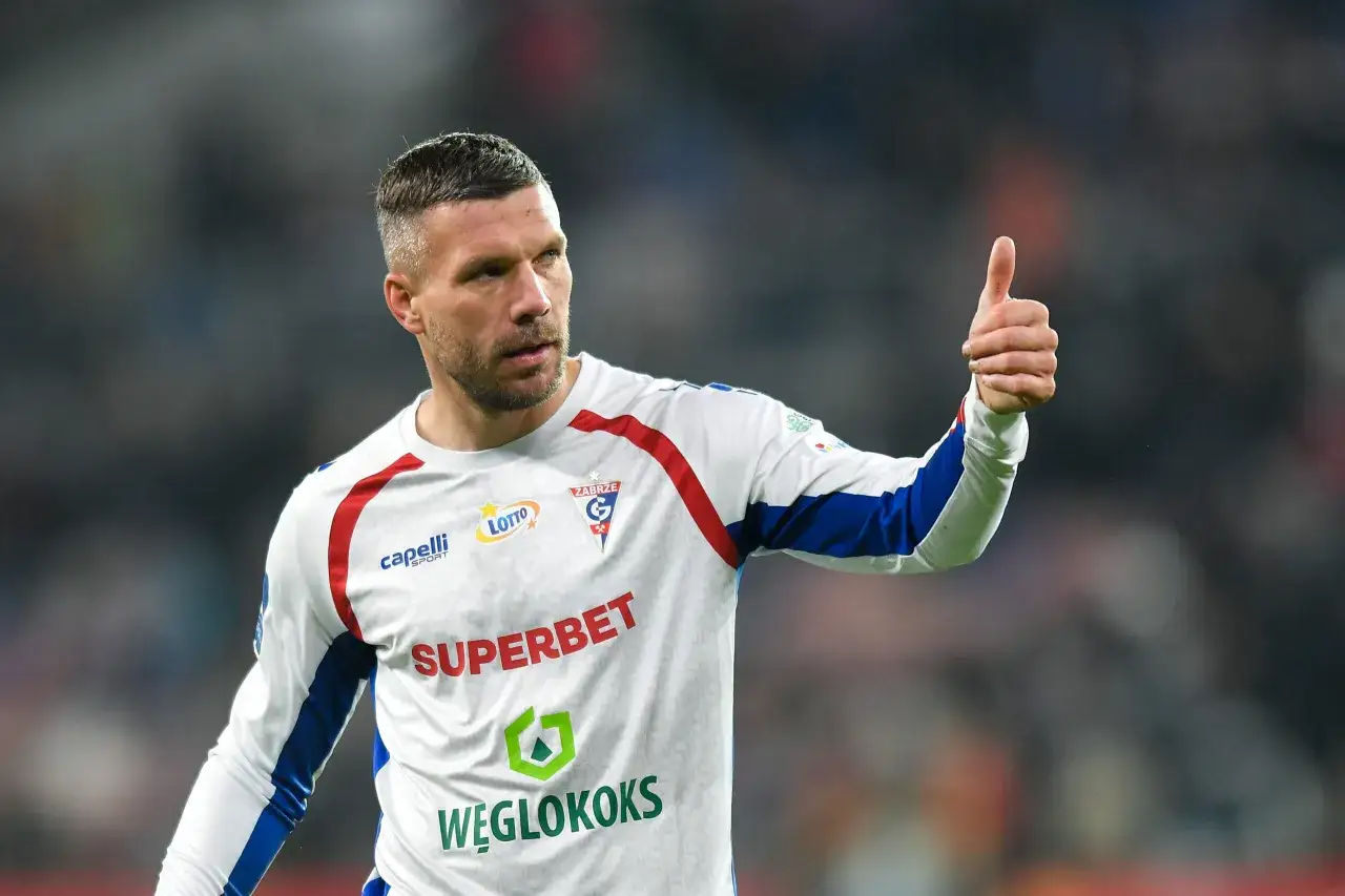Piłkarz w białej koszulce z czerwonymi i niebieskimi elementami pokazuje kciuk w górę. Mimo podolski wiek, wciąż w formie.