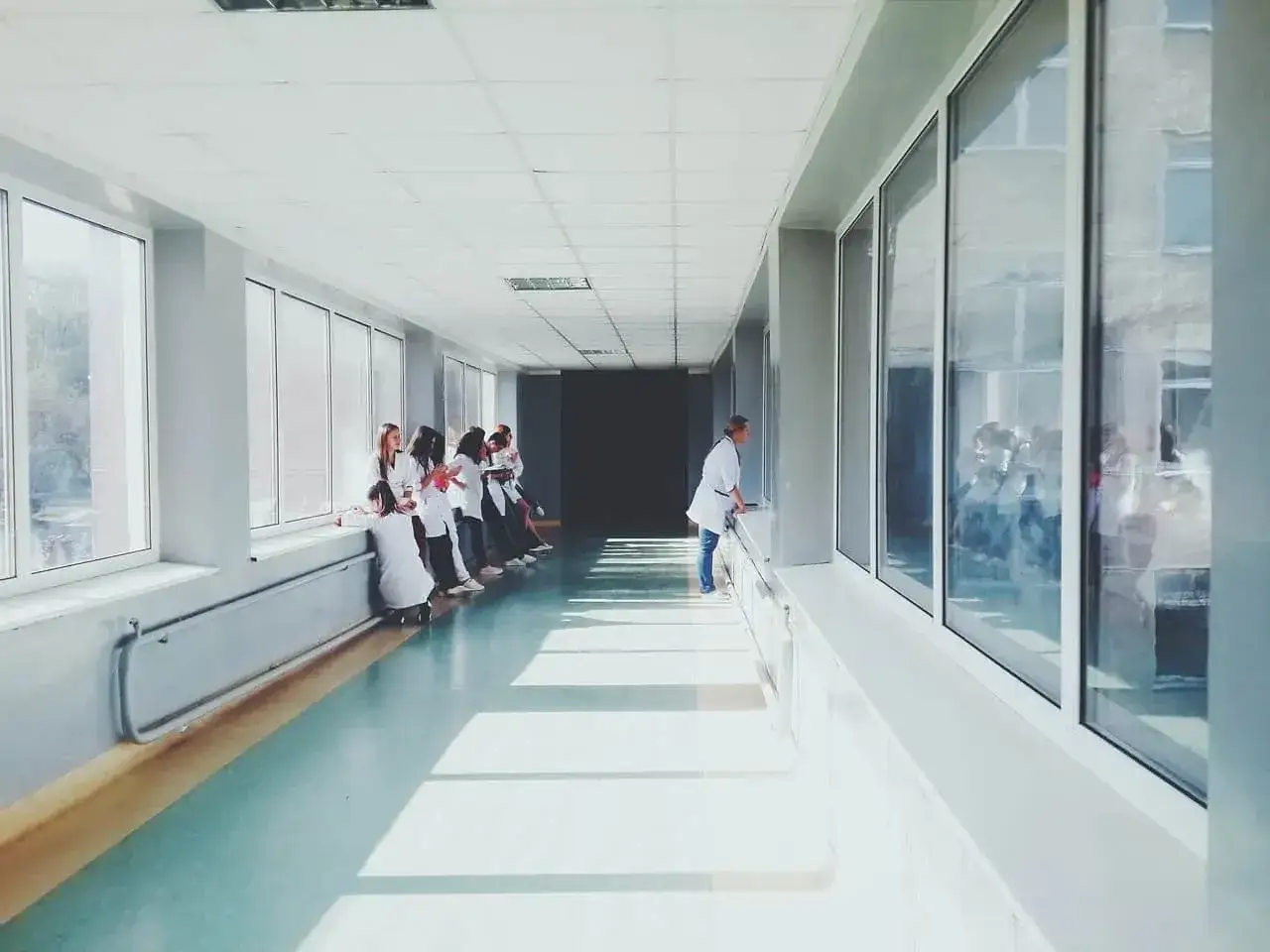 Grupa młodych ludzi w białych fartuchach, być może przyszli lekarze, siedzi na parapecie w długim korytarzu. Jeden z nich spogląda przez okno, jakby marząc o wolności, która może być daleka jak dożywotni pobyt w szpitalu psychiatrycznym.