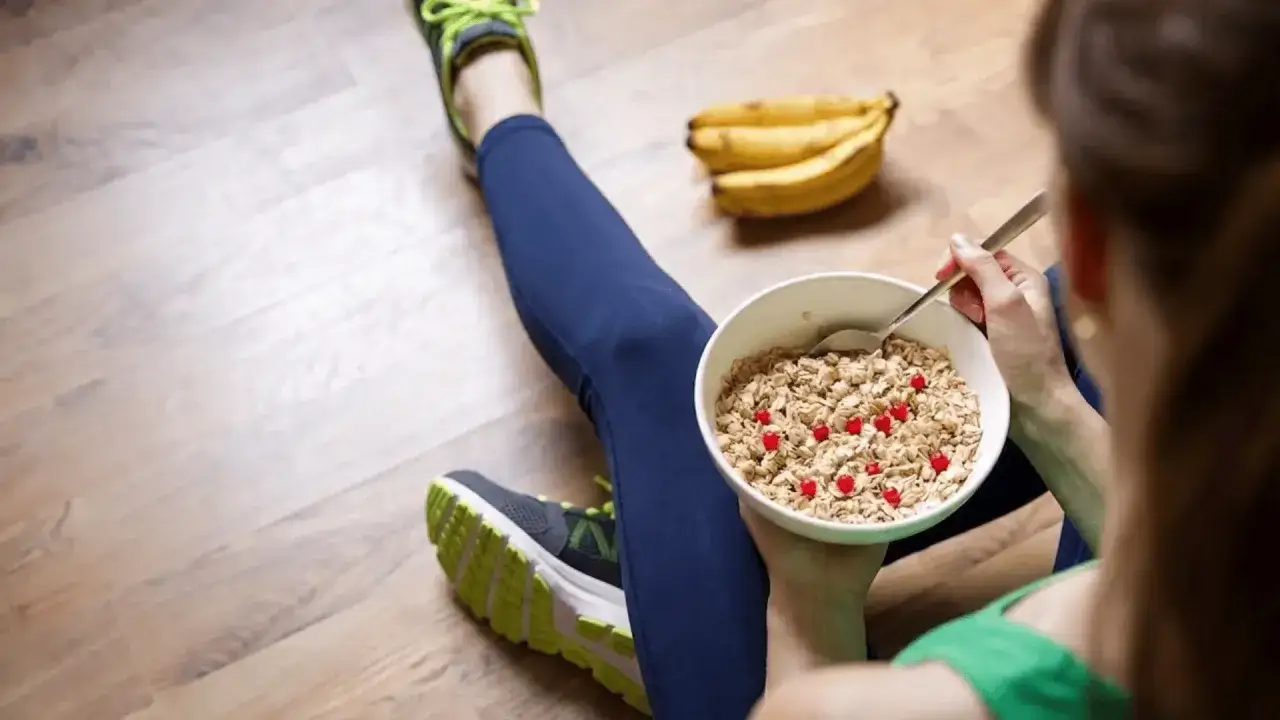 Una persona con abbigliamento sportivo mangia una ciotola di muesli con frutti rossi, con un grappolo di banana dopo allenamento sullo sfondo.