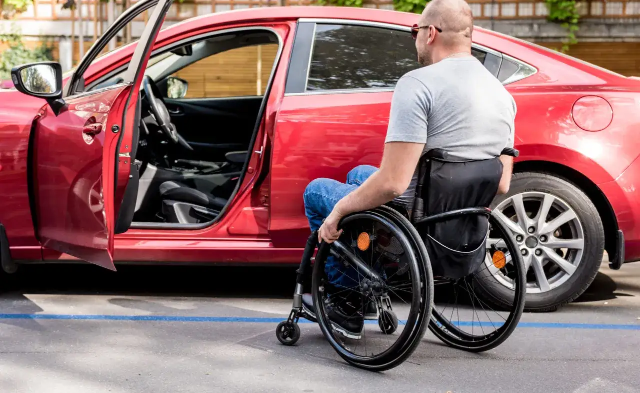 Uomo in sedia a rotelle vicino a una macchina rossa, con la portiera aperta. Il rigo e25 730 segna il parcheggio accessibile.