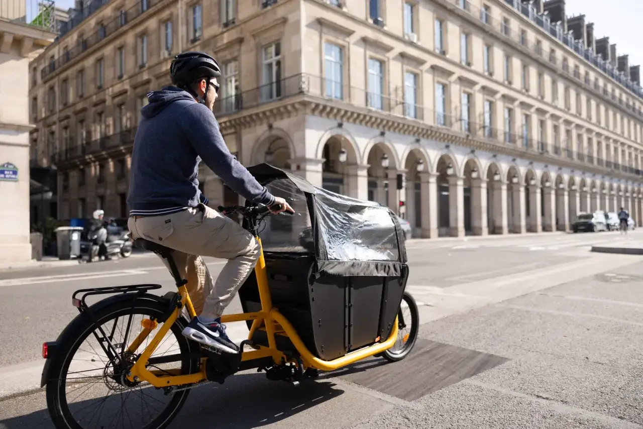 Un homme roule &agrave; Paris sur un v&eacute;lo cargo &eacute;lectrique jaune, son poids est bien r&eacute;parti pour une conduite stable.
