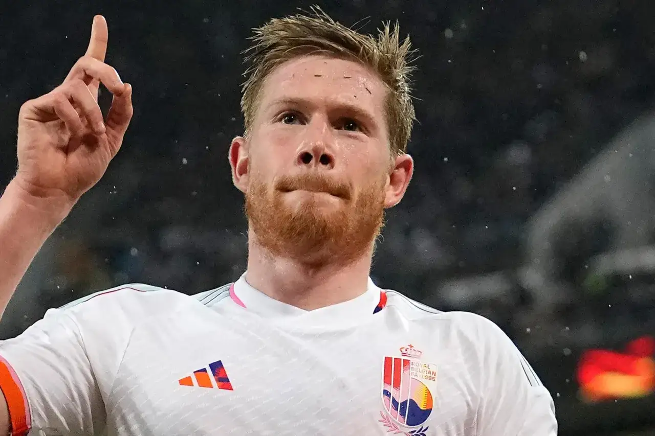 Kevin De Bruyne, w białej koszulce reprezentacji Belgii, unosi palec wskazujący, celebrując bramkę. Jego obecne drużyny to Manchester City i reprezentacja Belgii.