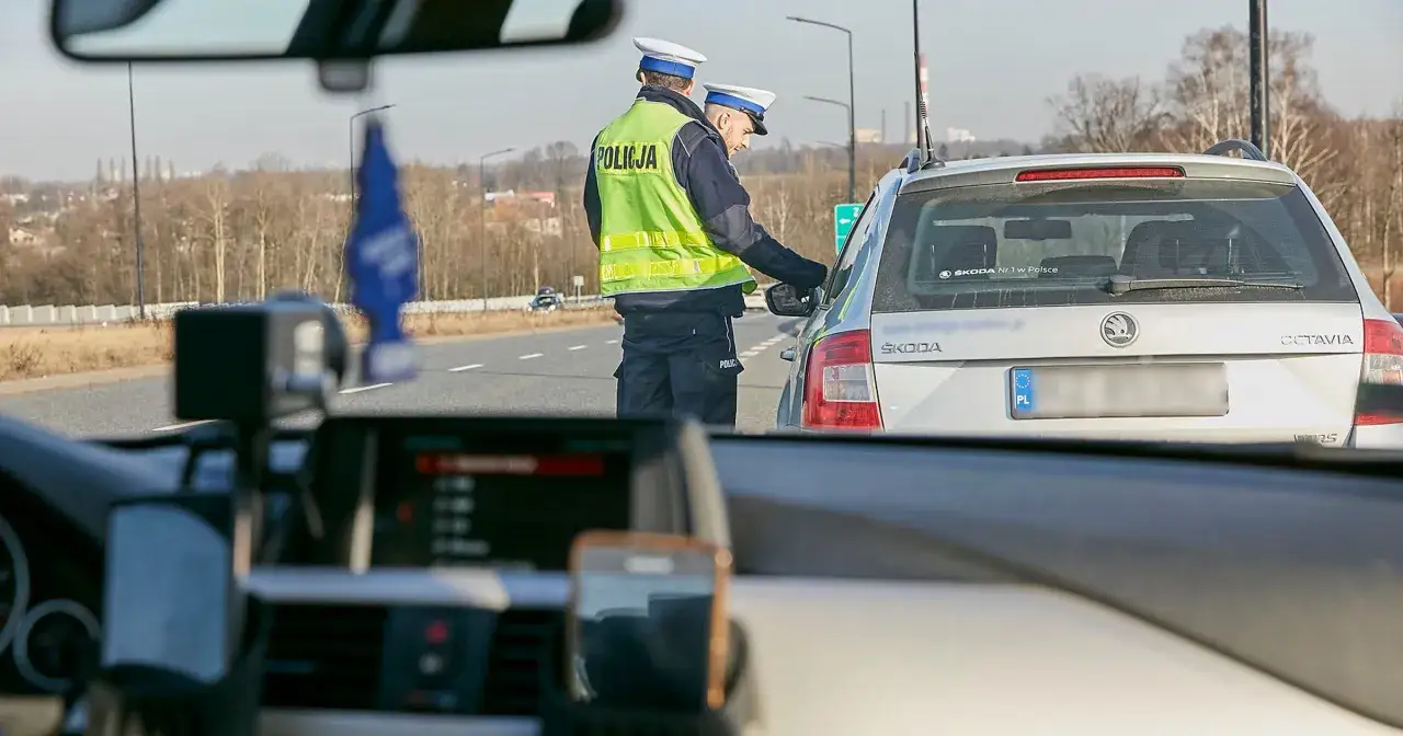 Policjanci w kamizelkach odblaskowych zatrzymali samochód. Jeden z nich bada go narkotestem, sprawdzając trzeźwość kierowcy.