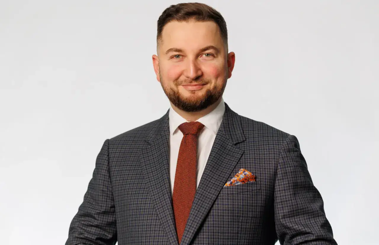 Mężczyzna w garniturze i krawacie, uśmiecha się do kamery.