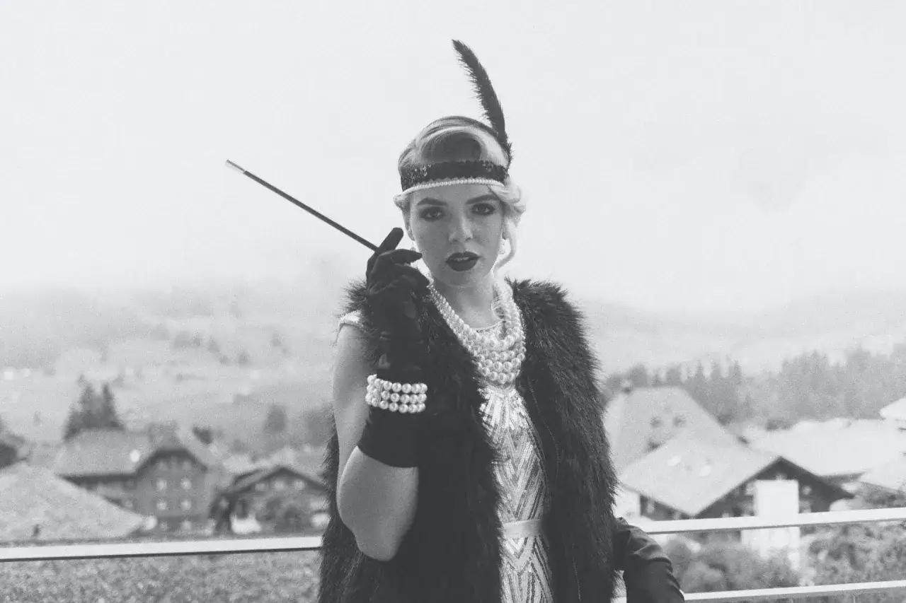 En kvinna i mode 1920-tal med fjäderpannband och pärlhalsband håller en cigarettmunstycke.