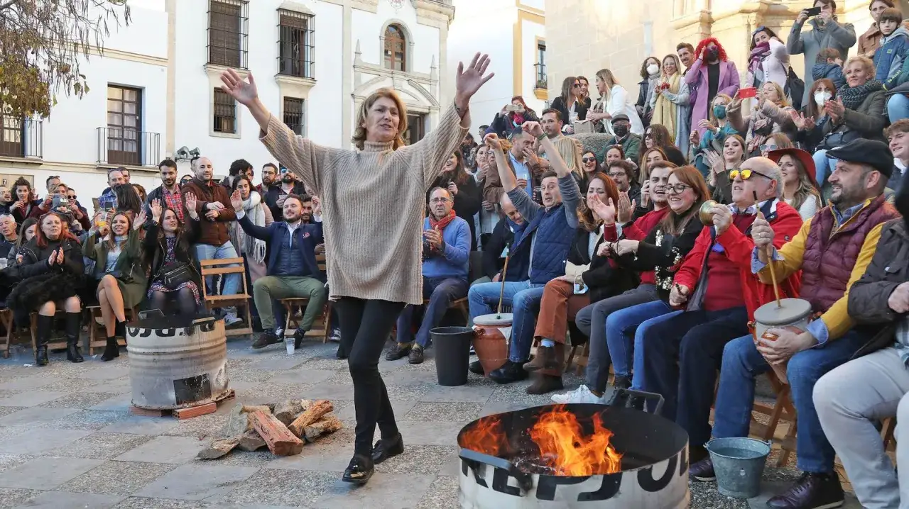 Una mujer baila animada mientras una multitud celebra las rumbas navideñas. El fuego crepita en un barril, creando un ambiente festivo.