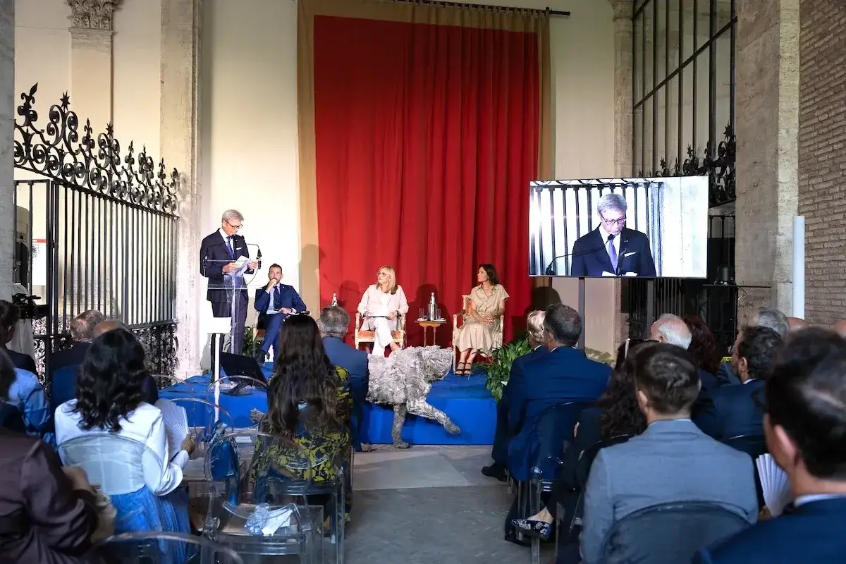 Un uomo parla a un podio durante un meeting per l'amicizia fra i popoli. Sullo schermo, un altro uomo parla.