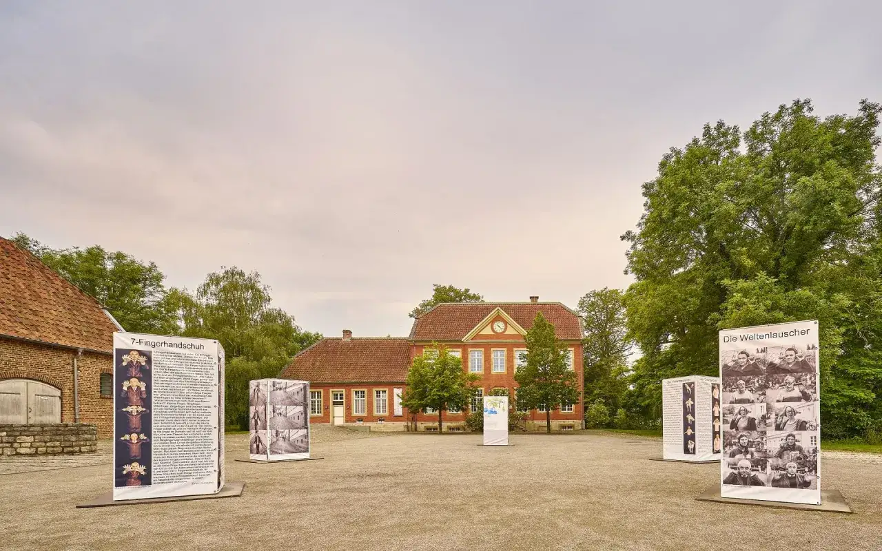 Das Haus Nottbeck mit Ausstellungen im Freien. Ein rotes Backsteingeb&auml;ude mit Ziegeldach und Uhr. Mehrere Informationstafeln stehen davor.