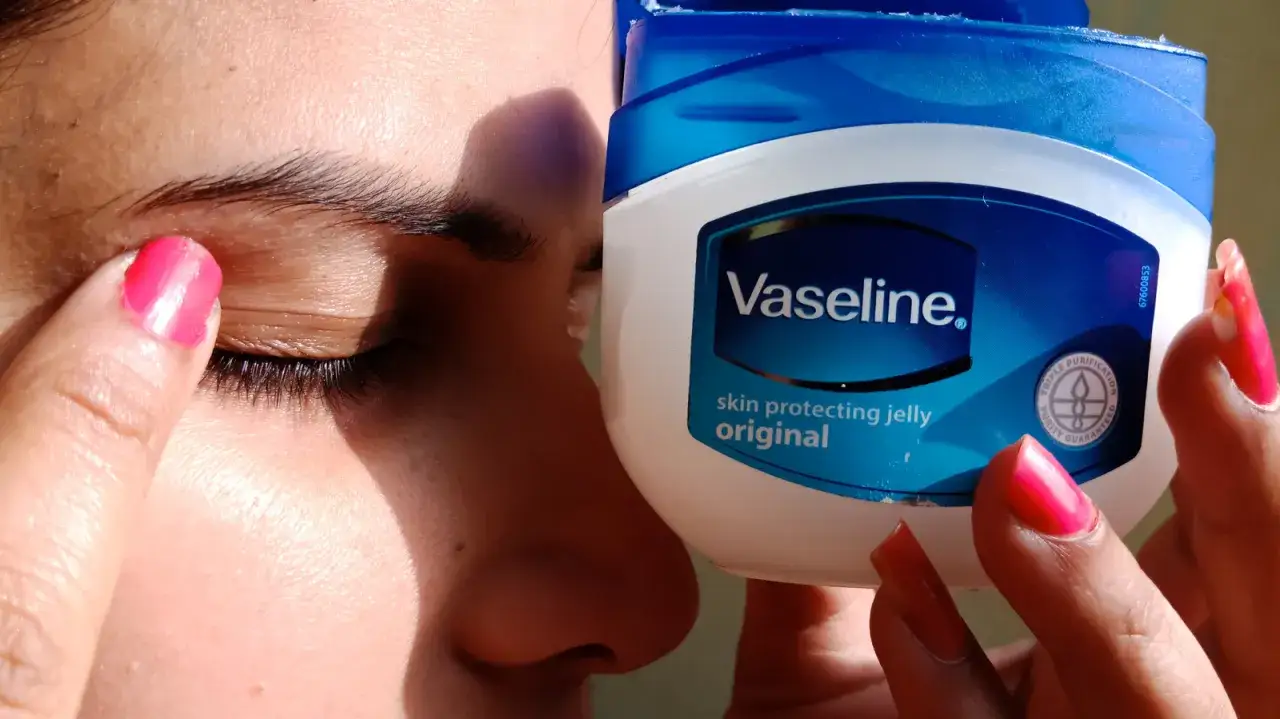 Palec z różowym lakierem nakłada wazelinę na powiekę. Obok słoiczek Vaseline.
