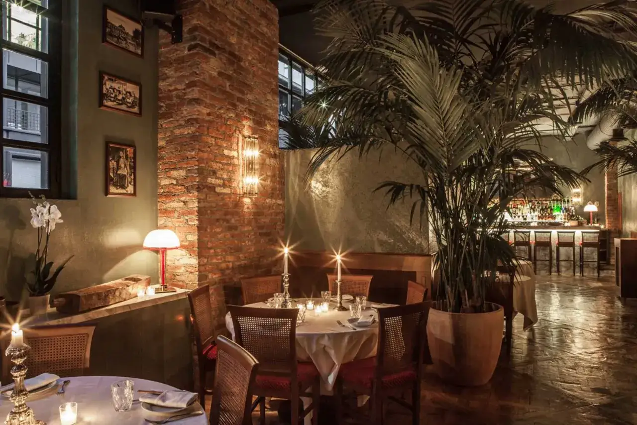 Atmosfera intima in uno dei **best restaurants in Milan**. Tavoli apparecchiati con candele, piante tropicali e un bar illuminato creano un ambiente accogliente.