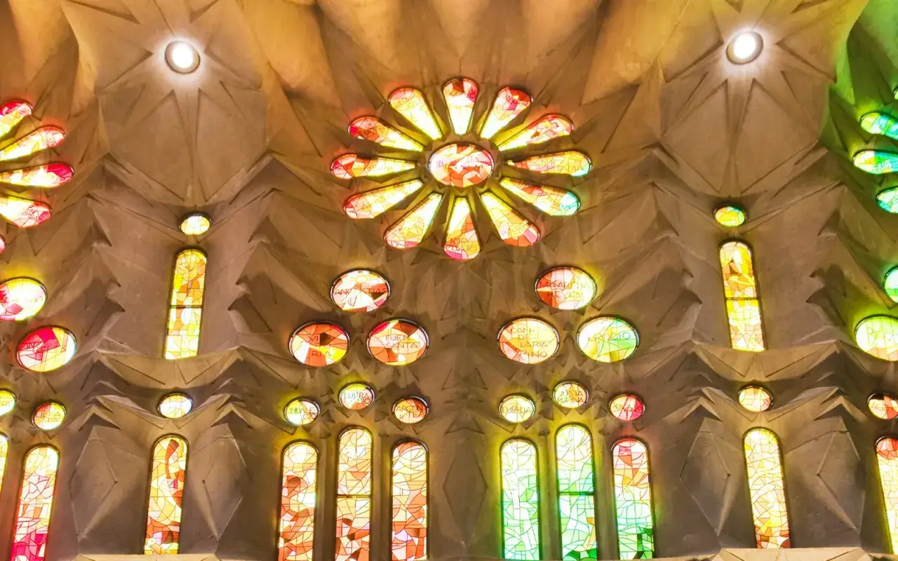 Wnętrze Sagrada Familia rozświetlone witrażami. Kolorowe światło przechodzi przez okna, tworząc mozaikę barw na kamiennych ścianach i sklepieniu.