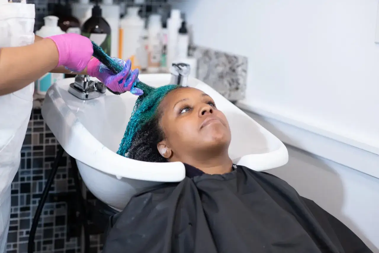 Une personne en gants roses applique un soin bleu turquoise sur les cheveux d'une femme dans une bac de lavage. C'est une &eacute;tape cl&eacute; pour savoir comment bien rincer ses cheveux.