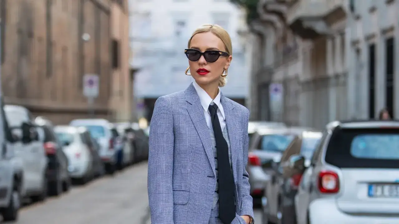 Mujer tomboy con traje a cuadros, camisa blanca y corbata negra, gafas de sol y labios rojos.