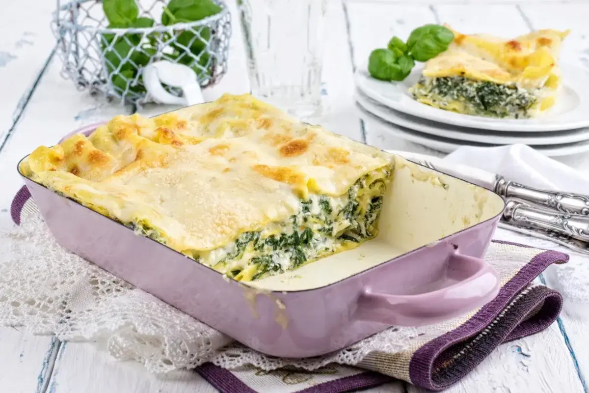Herzhafte Spinat-Lasagne in einer lila Auflaufform, goldbraun überbacken. Ein Stück auf einem Teller daneben.
