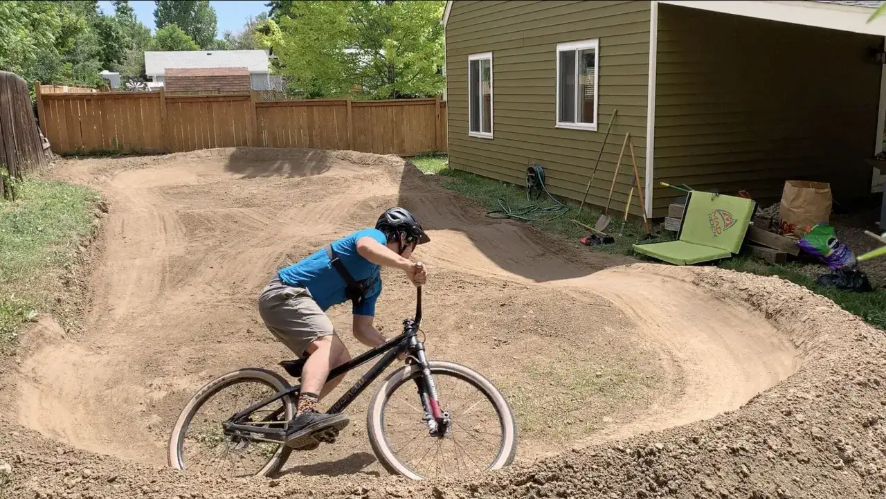 Chłopiec na rowerze testuje nowy **pump track** w ogrodzie. W tle dom i narzędzia ogrodnicze.