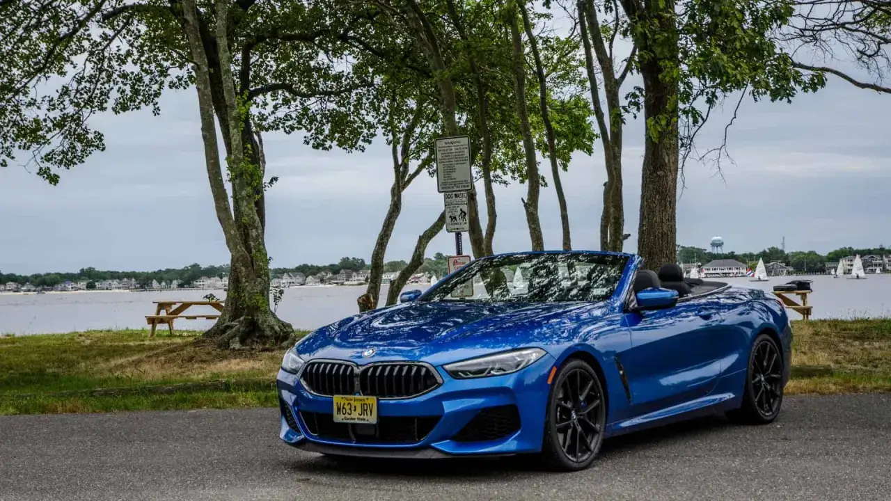 Błękitny kabriolet BMW M850i zaparkowany nad jeziorem, z drzewami i domami w tle.