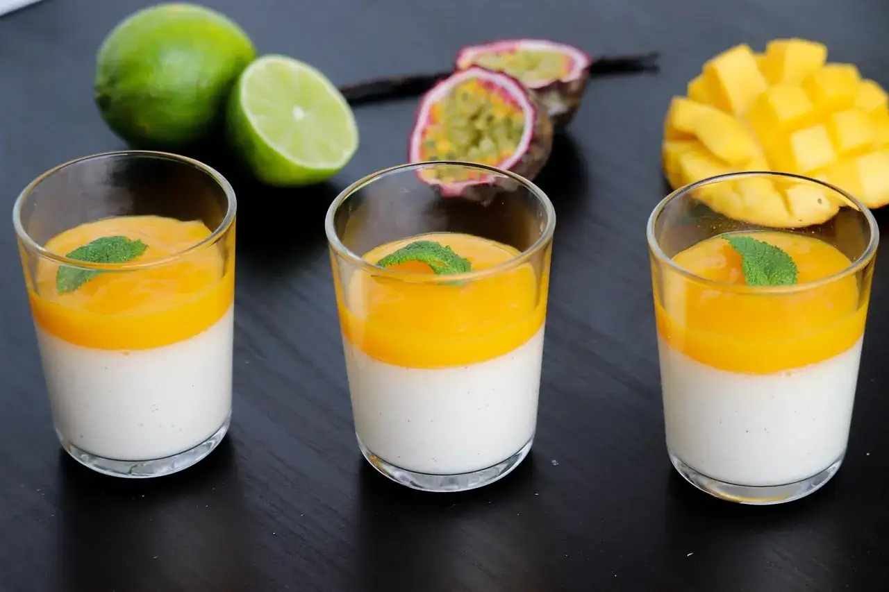 Trois panna cotta mangue, garnies de menthe, avec des fruits frais en arrière-plan.