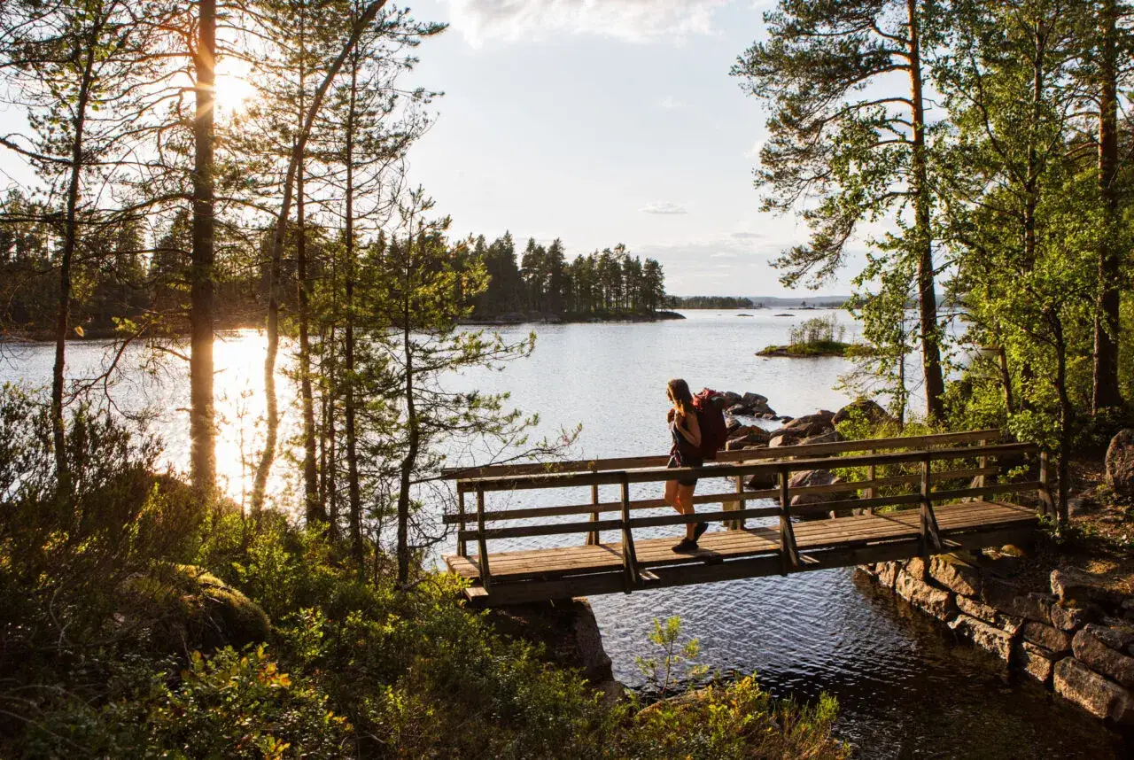 En person med ryggsäck korsar en träbro under en glaskogen vandring. Solen skiner genom träden vid en sjö.