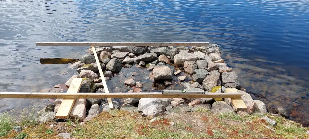 Arbete pågår för att bygga brygga vid en sjö. Stenar och träbjälkar ligger redo vid vattenbrynet.