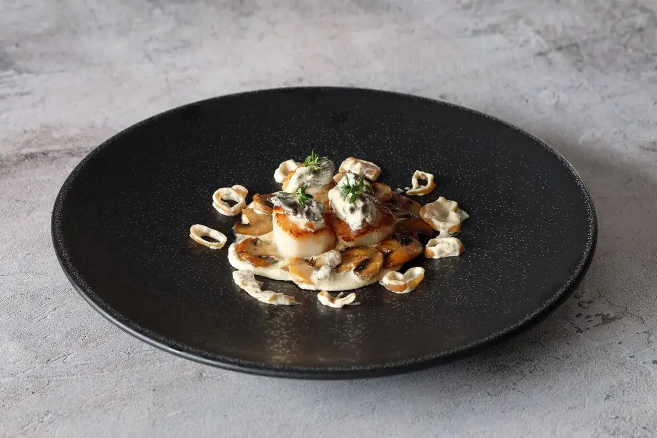 Coquilles Saint-Jacques aux morilles et leur sauce au vin blanc, garnies de thym frais, sur une assiette noire.