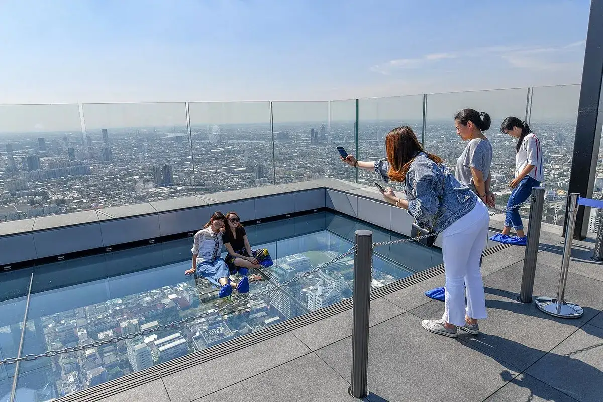 Besucher genie&szlig;en den atemberaubenden Blick vom Mahanakhon Skywalk. Zwei Frauen sitzen auf dem Glasboden, w&auml;hrend eine andere Fotos macht.