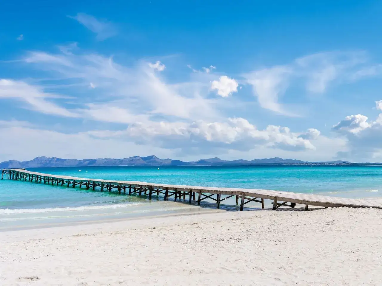 Holzsteg im türkisfarbenen Meer, Blick auf die Berge im mallorcas norden. Weißer Sandstrand, blauer Himmel mit Wolken.