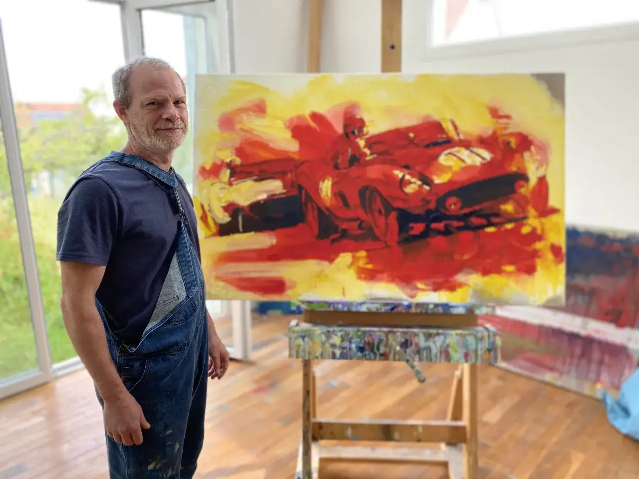 Un artiste peintre auto entrepreneur pose devant sa toile vibrante d'une voiture de course rouge.