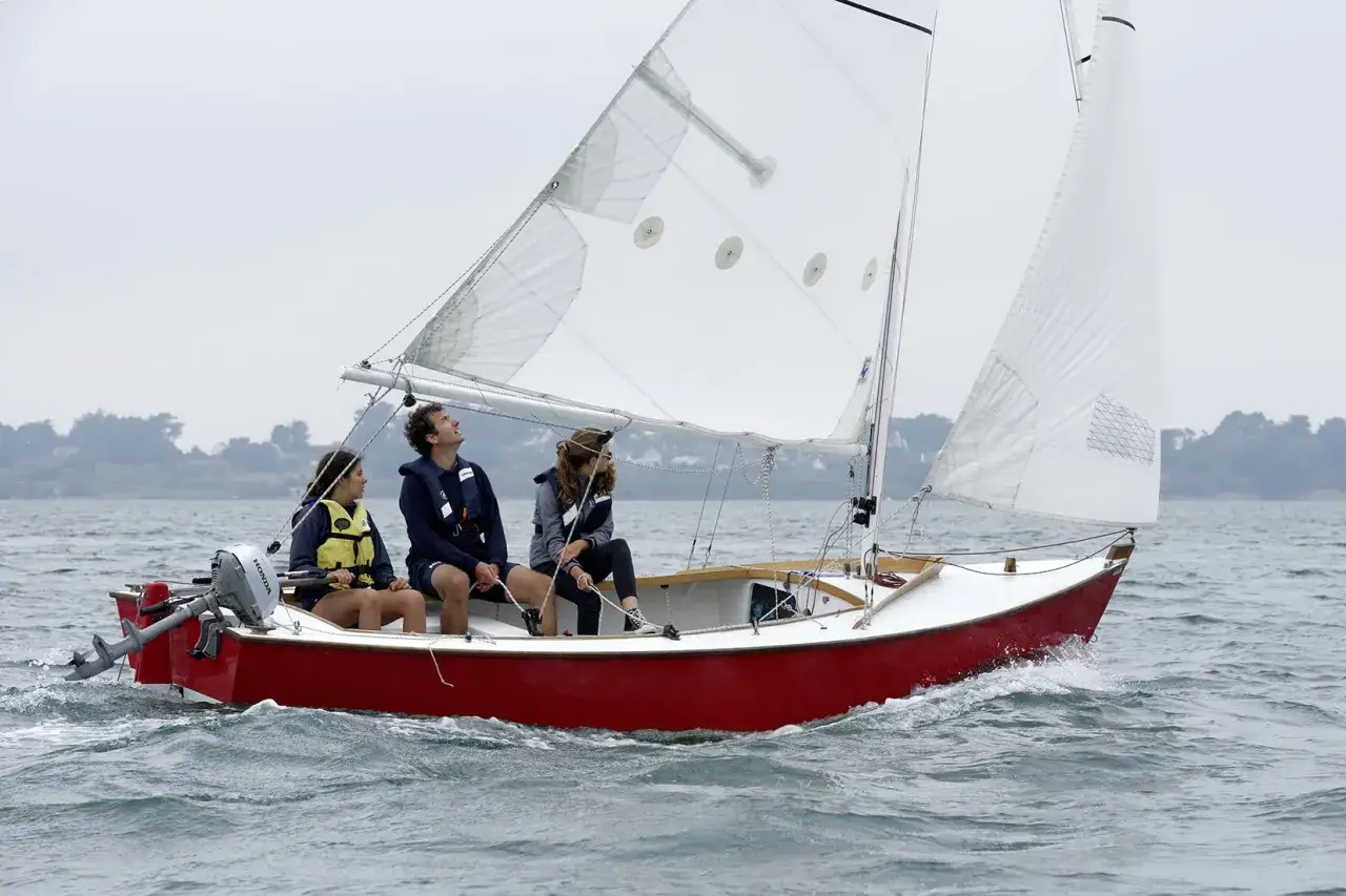 Trois personnes profitent d'une sortie en mer sur un petit voilier 2 places, le vent gonflant les voiles blanches.
