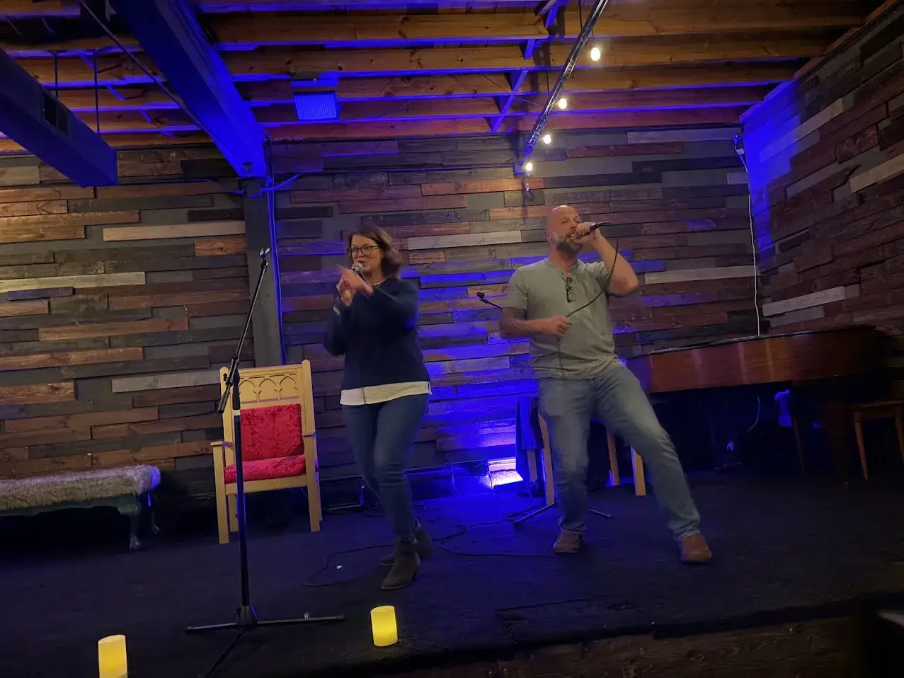 Ein Mann und eine Frau singen im Duett, vielleicht die **top 100 karaoke songs**. Sie stehen auf einer Bühne mit Holzwänden und blauer Beleuchtung.