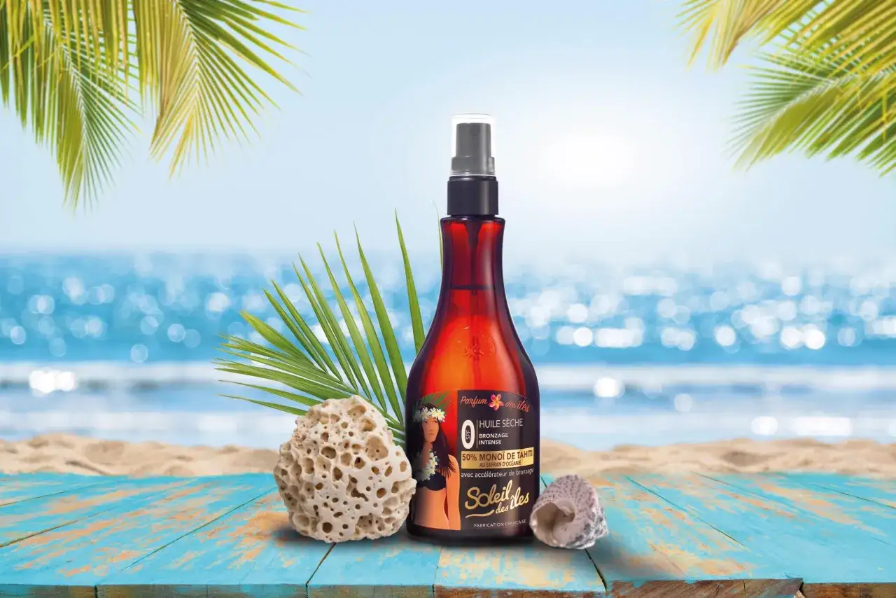 Flacon d'huile s&egrave;che bronzante au monoi de Tahiti, sur une plage de sable fin, avec des palmiers et la mer en arri&egrave;re-plan.