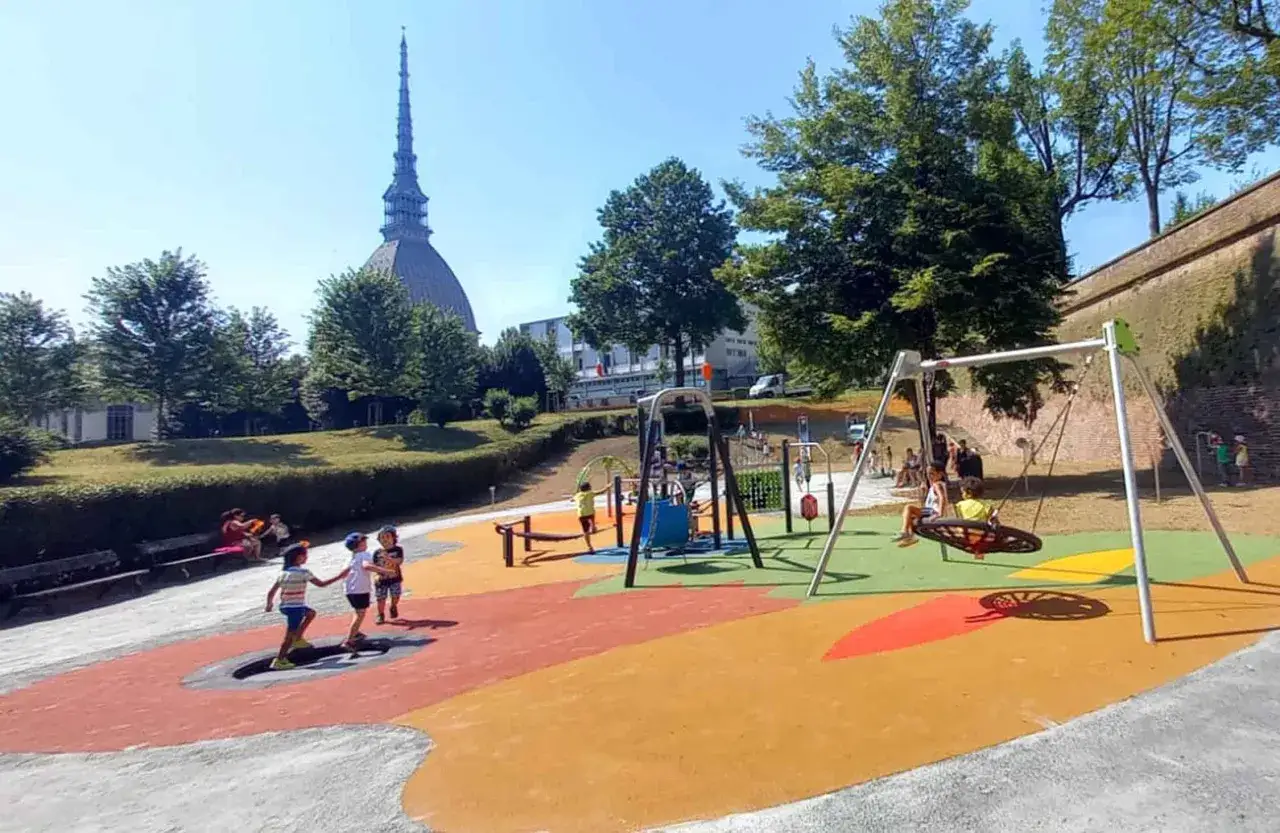 Un parco giochi colorato nel **Piemonte con bambini** che si divertono sull'altalena e sui giochi. In lontananza, la Mole Antonelliana.