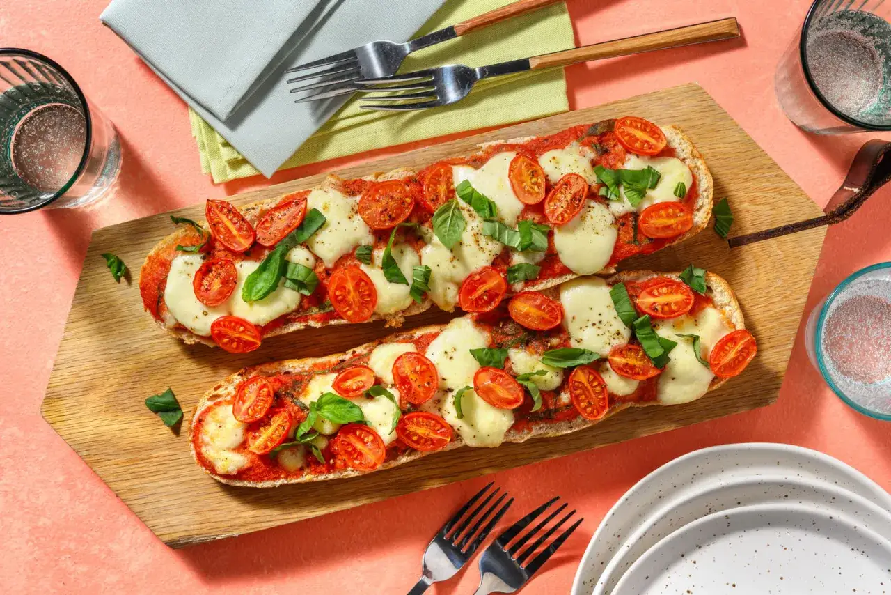 Deux baguettes garnies de sauce tomate, mozzarella fondue, tomates cerises et basilic, prêtes pour une recette de bruschetta au four.