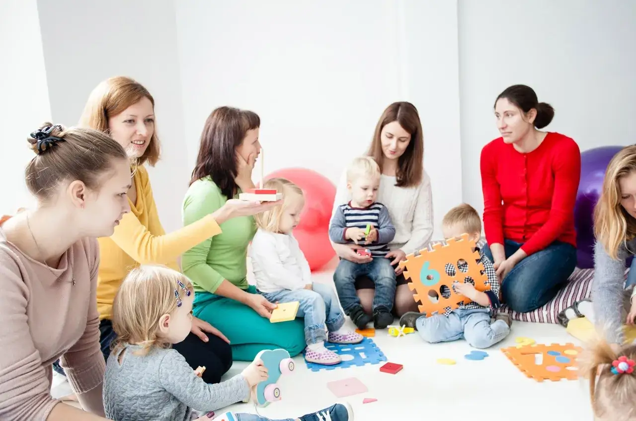 Grupa dzieci i dorosłych uczestniczy w zajęciach, które przypominają terapię pedagogiczną. Dzieci bawią się klockami i zabawkami edukacyjnymi.