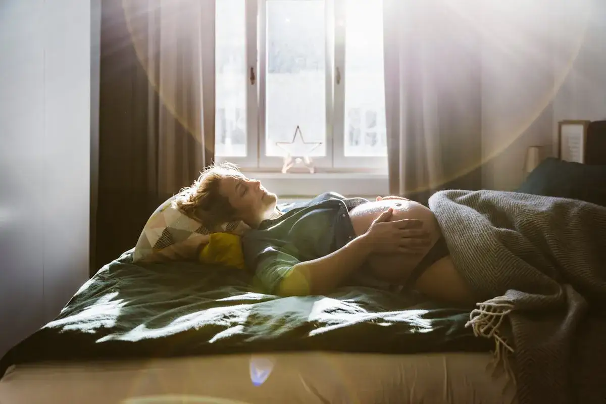 Femme enceinte, le ventre à l'air, repose sur un lit. La lumière du soleil filtre à travers la fenêtre, créant une atmosphère douce. Elle semble lutter contre l'insomnie début grossesse.
