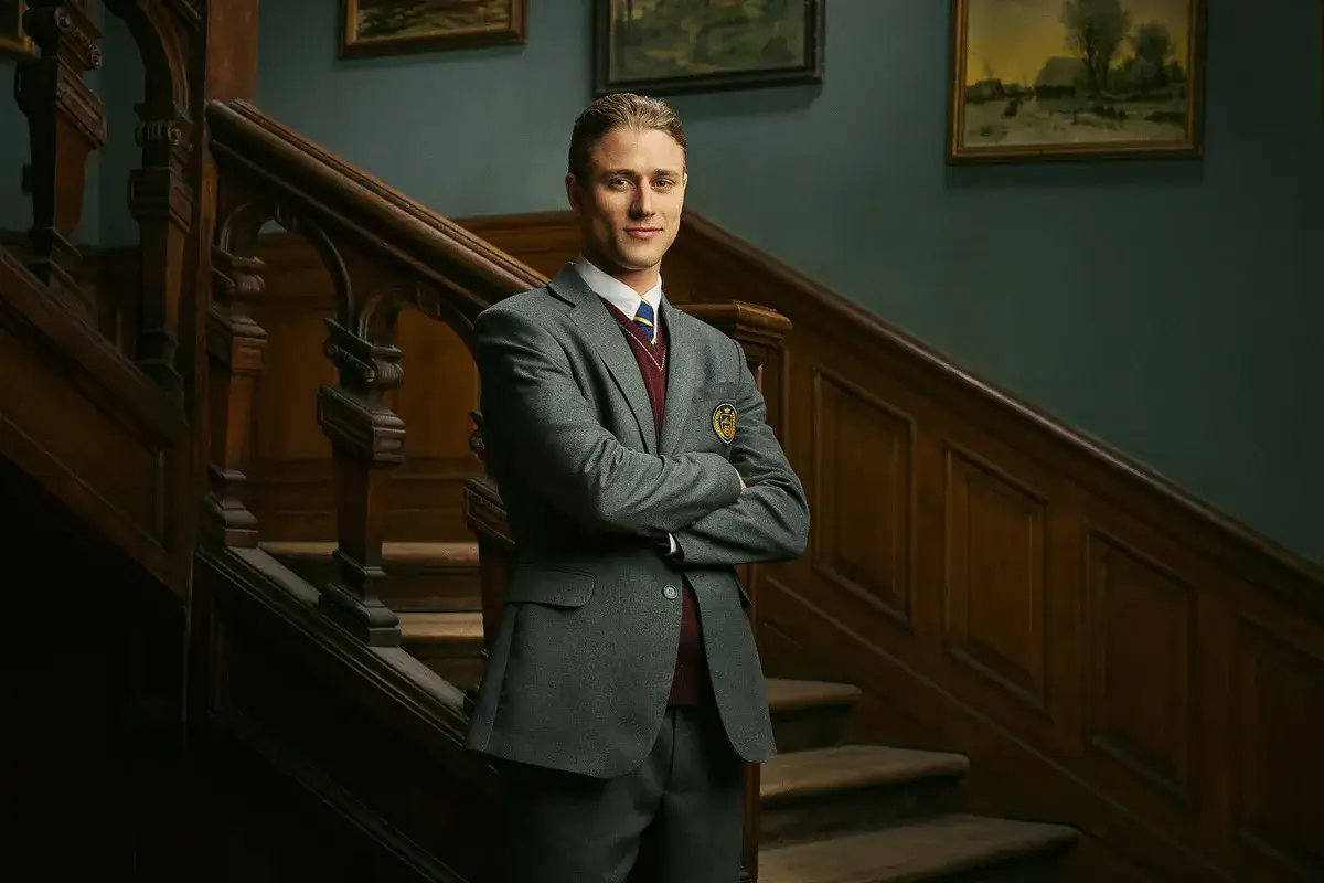En stilig ung man i skoluniform står med armarna i kors på en trä trappa. Han är en av karaktärerna i rollistan i ondskan (tv-serie).