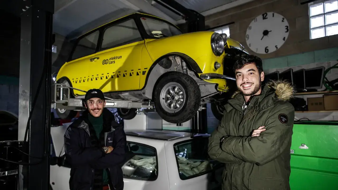 Dos hombres sonr&iacute;en junto a un Mini amarillo en un taller. El coche est&aacute; elevado, mostrando su parte inferior. Este es un ejemplo de retrofit, una transformaci&oacute;n.