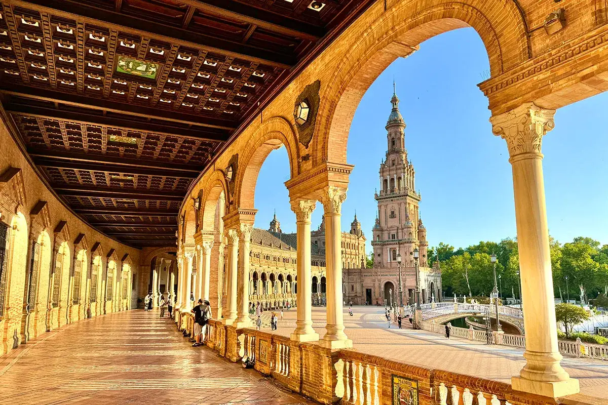 Sonnenschein beleuchtet die Arkaden der Plaza de Espa&ntilde;a, ein Wahrzeichen Sevilles. Ein Turm ragt im Hintergrund auf.