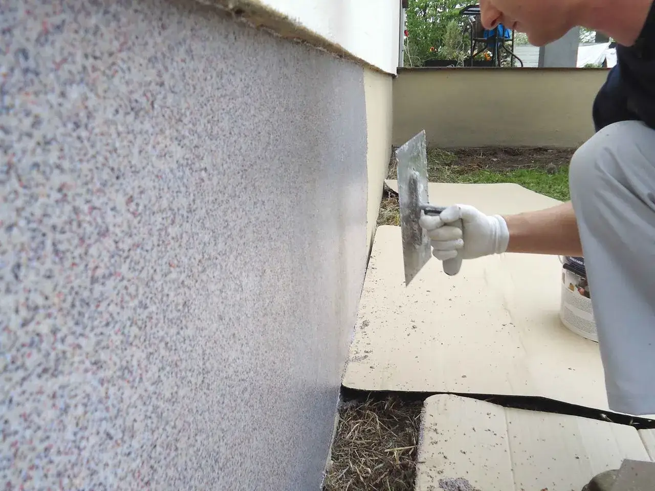 Nakładanie tynku mozaikowego na ścianę za pomocą kielni. W tle widać pojemnik z produktem.
