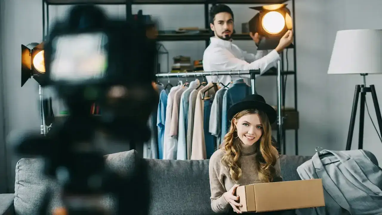 Kobieta z pudełkiem i mężczyzna z lampą przygotowują się do sesji live commerce. W tle wieszaki z ubraniami.