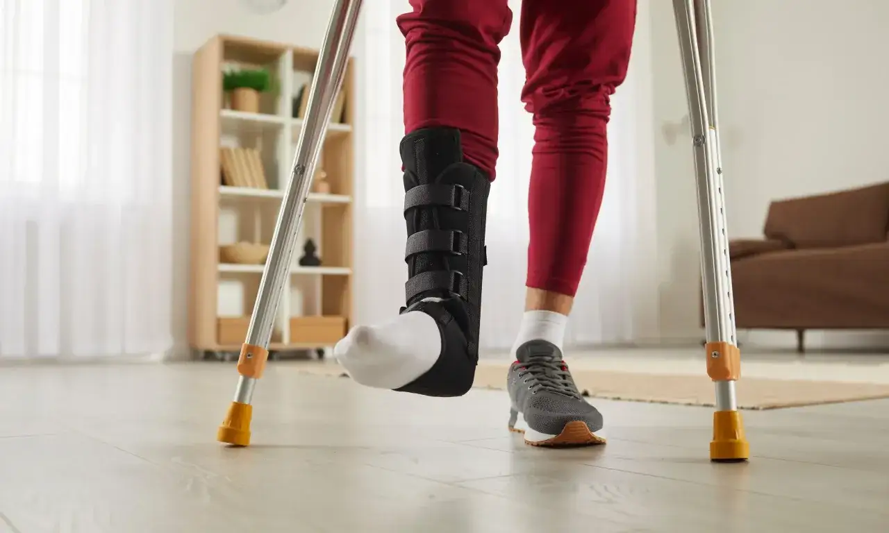 Osoba w ortezie na stawie skokowym, wspierając się na kulach, stawia pierwsze kroki po operacji.