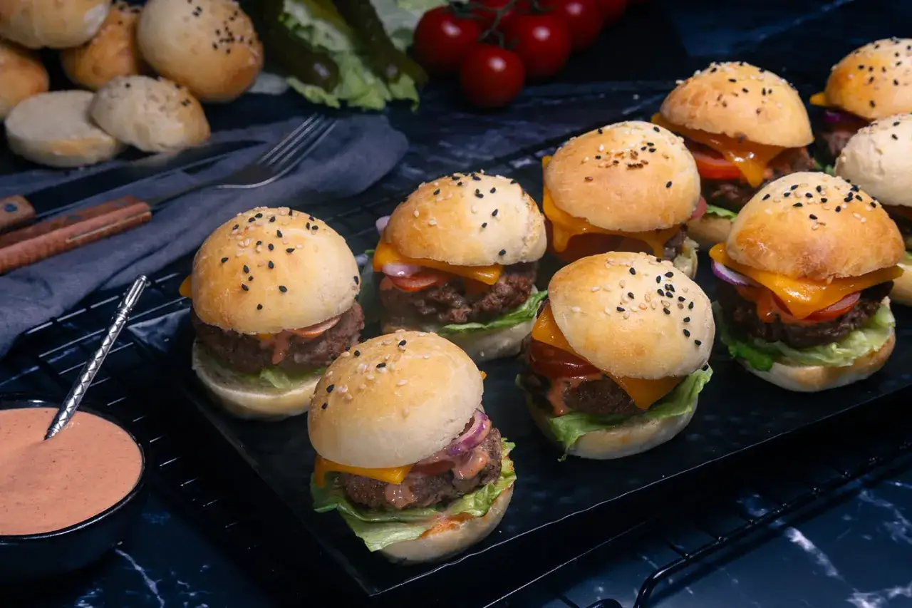 Mehrere Mini Burger mit Käse, Salat und Tomaten auf einem Rost. Daneben eine Schale mit Sauce.