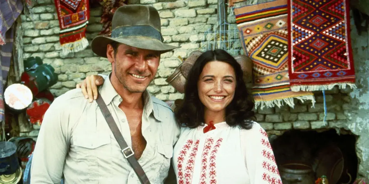 Indiana Jones i Marion Ravenwood, czyli poszukiwacze zaginionej arki obsada, uśmiechają się na tle kolorowych dywanów.