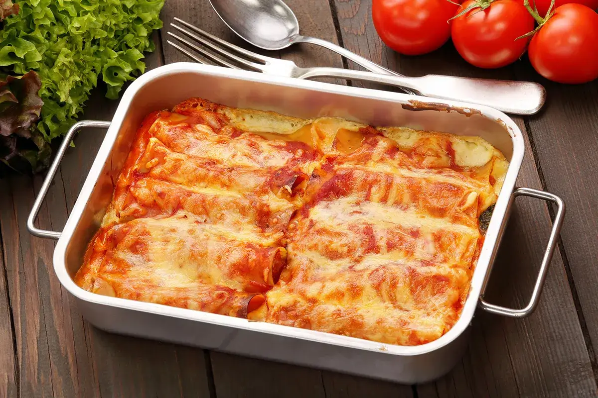 Canelones de at&uacute;n gratinados en una bandeja met&aacute;lica, acompa&ntilde;ados de tomates y lechuga fresca.