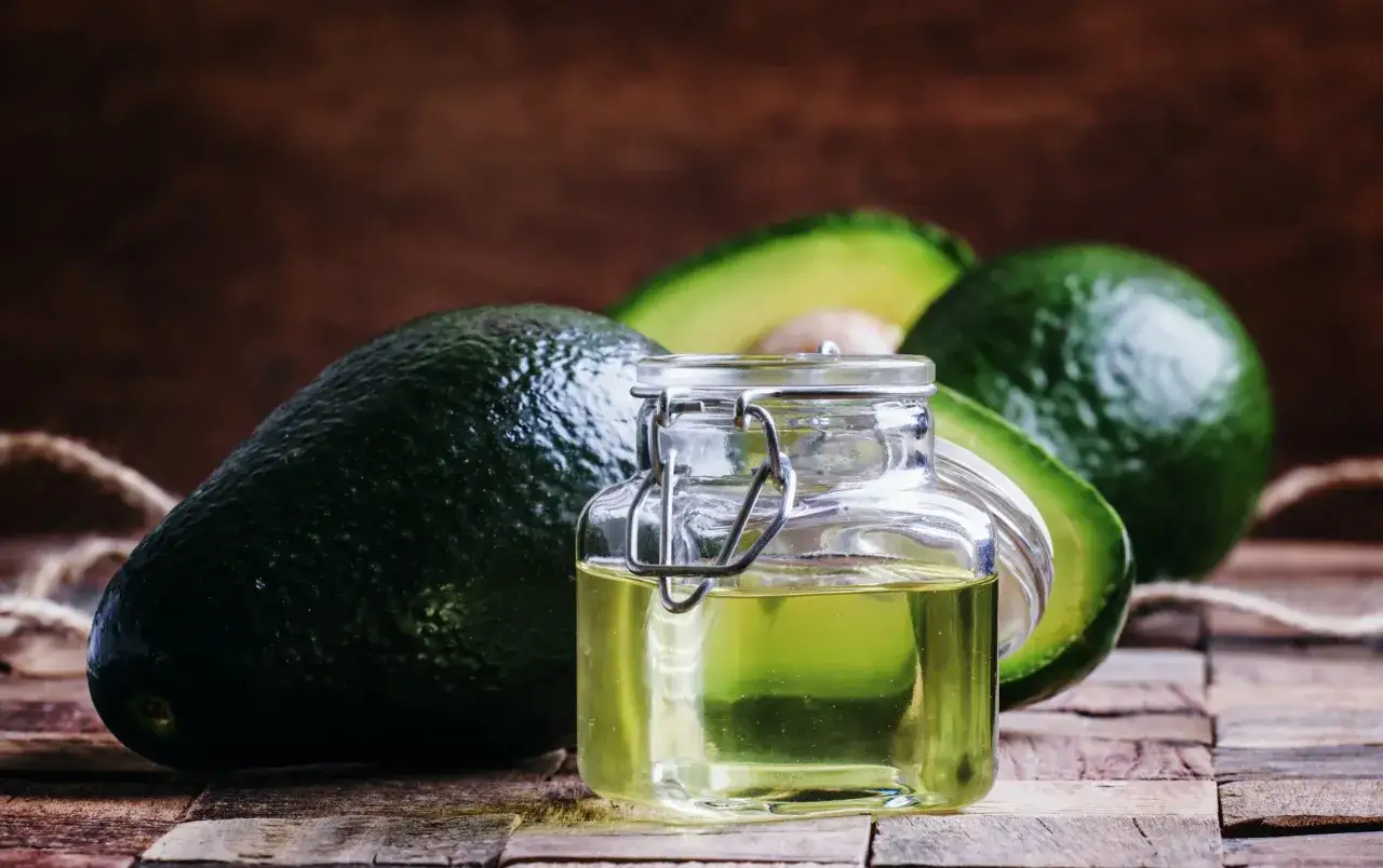 Un frasco de vidrio con aceite de aguacate para la piel, junto a aguacates frescos sobre madera r&uacute;stica.