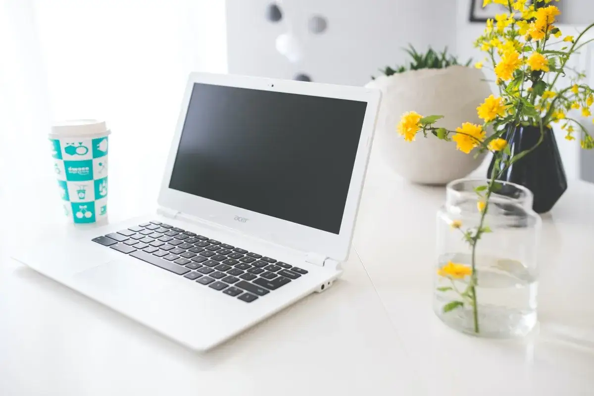 Biały chromebook, kubek z napisem i żółte kwiaty tworzą przytulne miejsce do pracy.