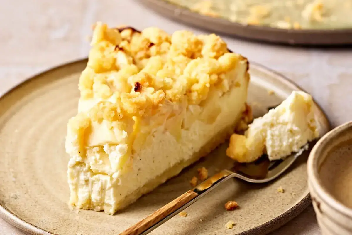 Ein Stück Apfel Quark Kuchen mit Streuseln auf einem Teller. Ein Gabelstück liegt daneben.